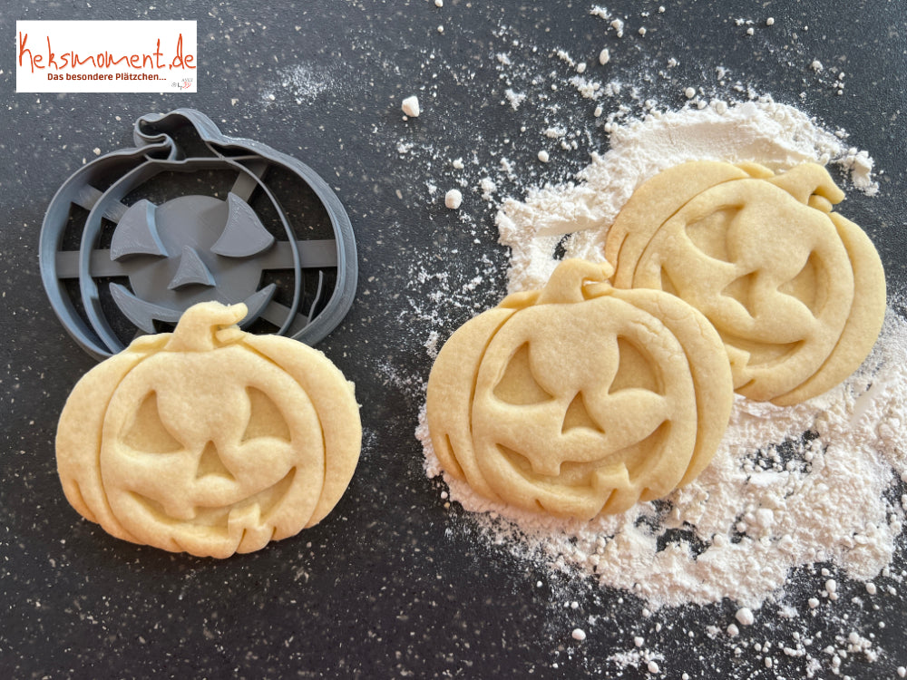 Halloweenkürbis-Ausstecher neben drei ausgestochenen Kürbismotiven mit grinsendem Gesicht auf bemehlter Arbeitsfläche.