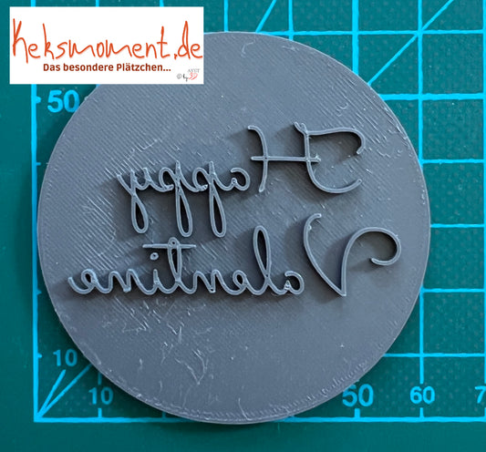 Runder Stempel mit spiegelverkehrtem Schriftzug Happy Valentine auf einer Schneidematte mit Millimeterskala.